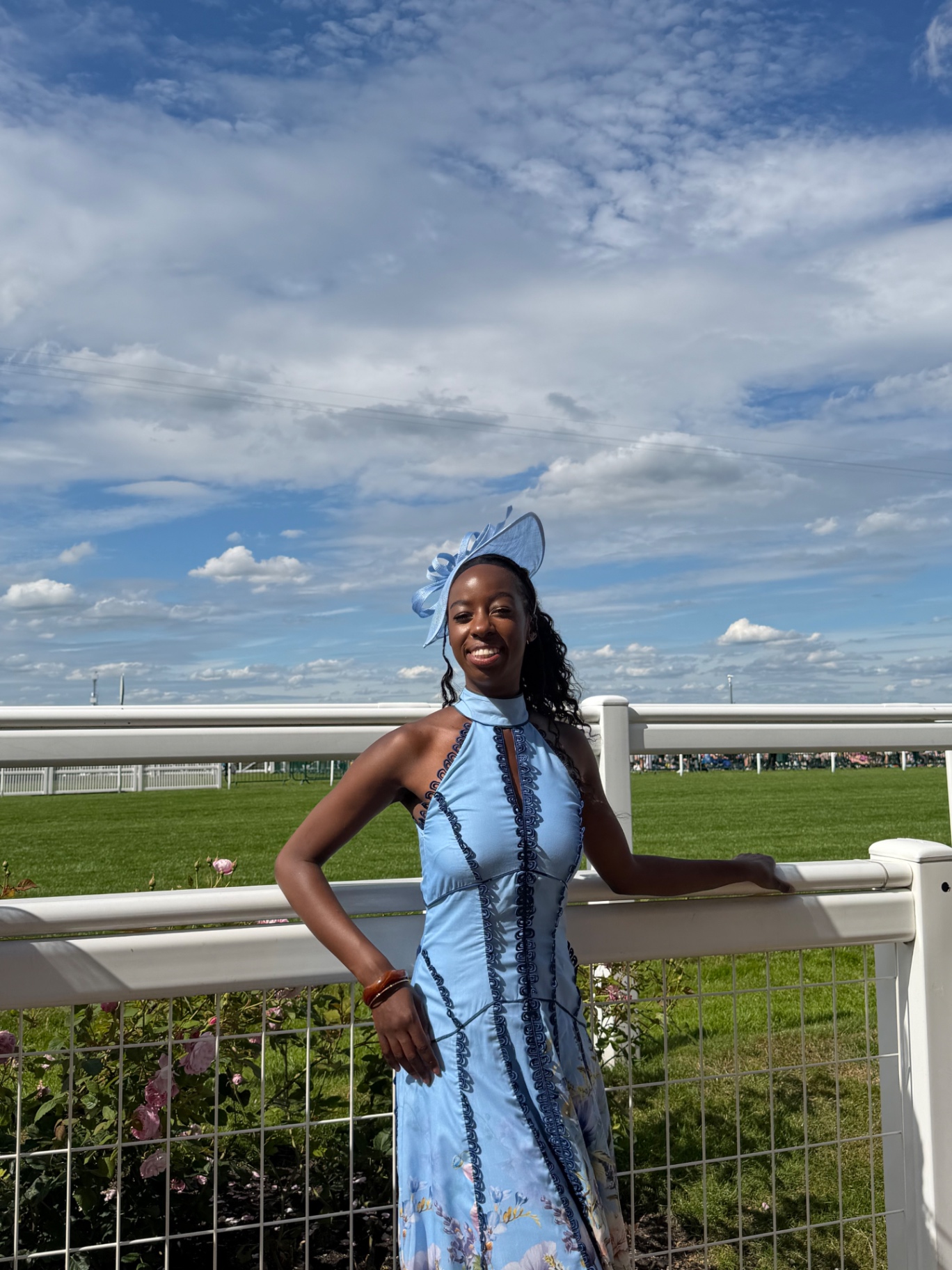 A member in a blue halter dress and fascinator by the racecourse rail