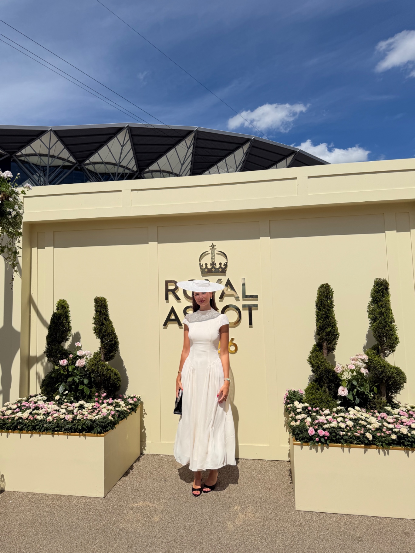 A member in a white dress and boater hat at the Royal Ascot sign