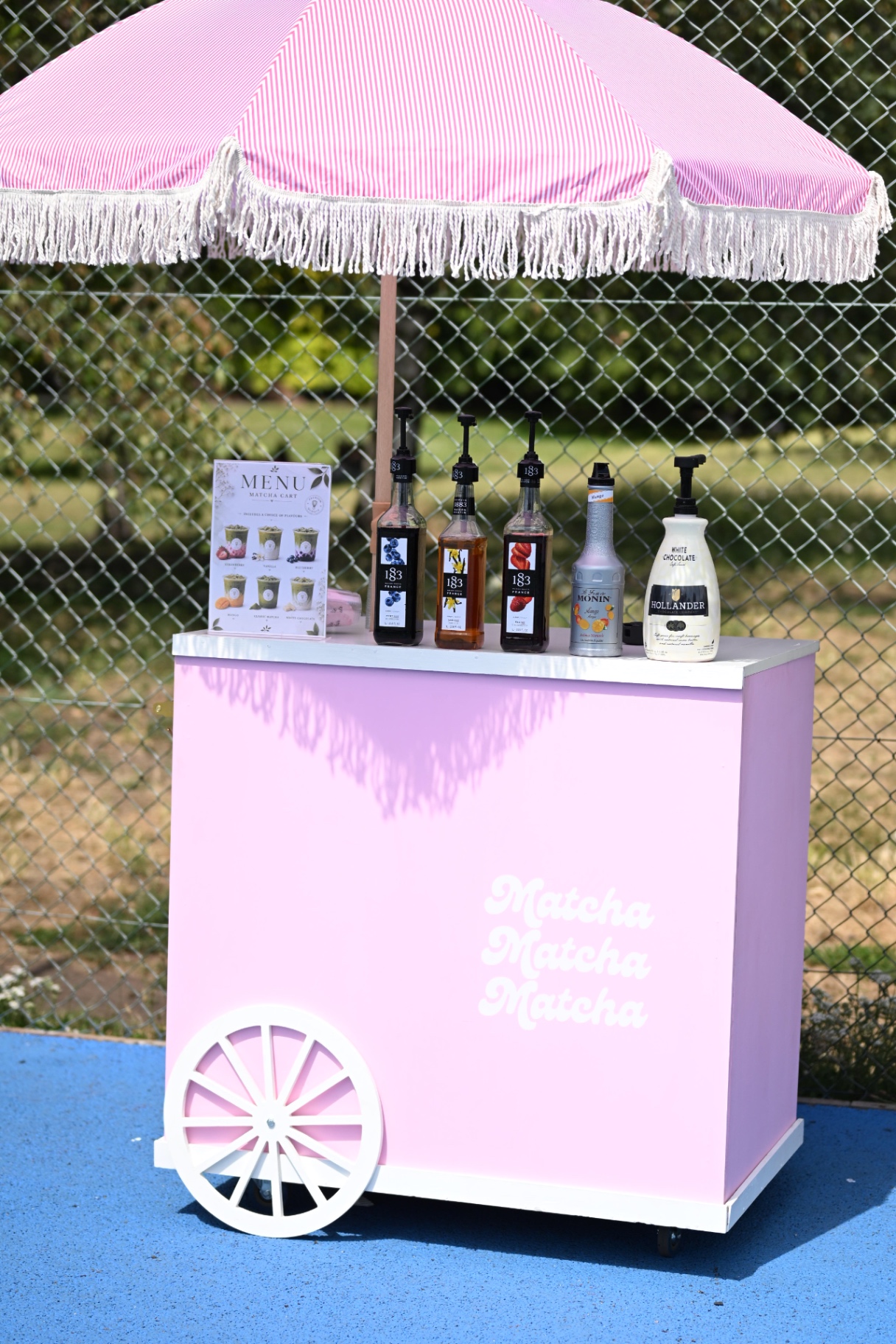 The pink La Vie Mayfair matcha cart with syrups under a fringed parasol