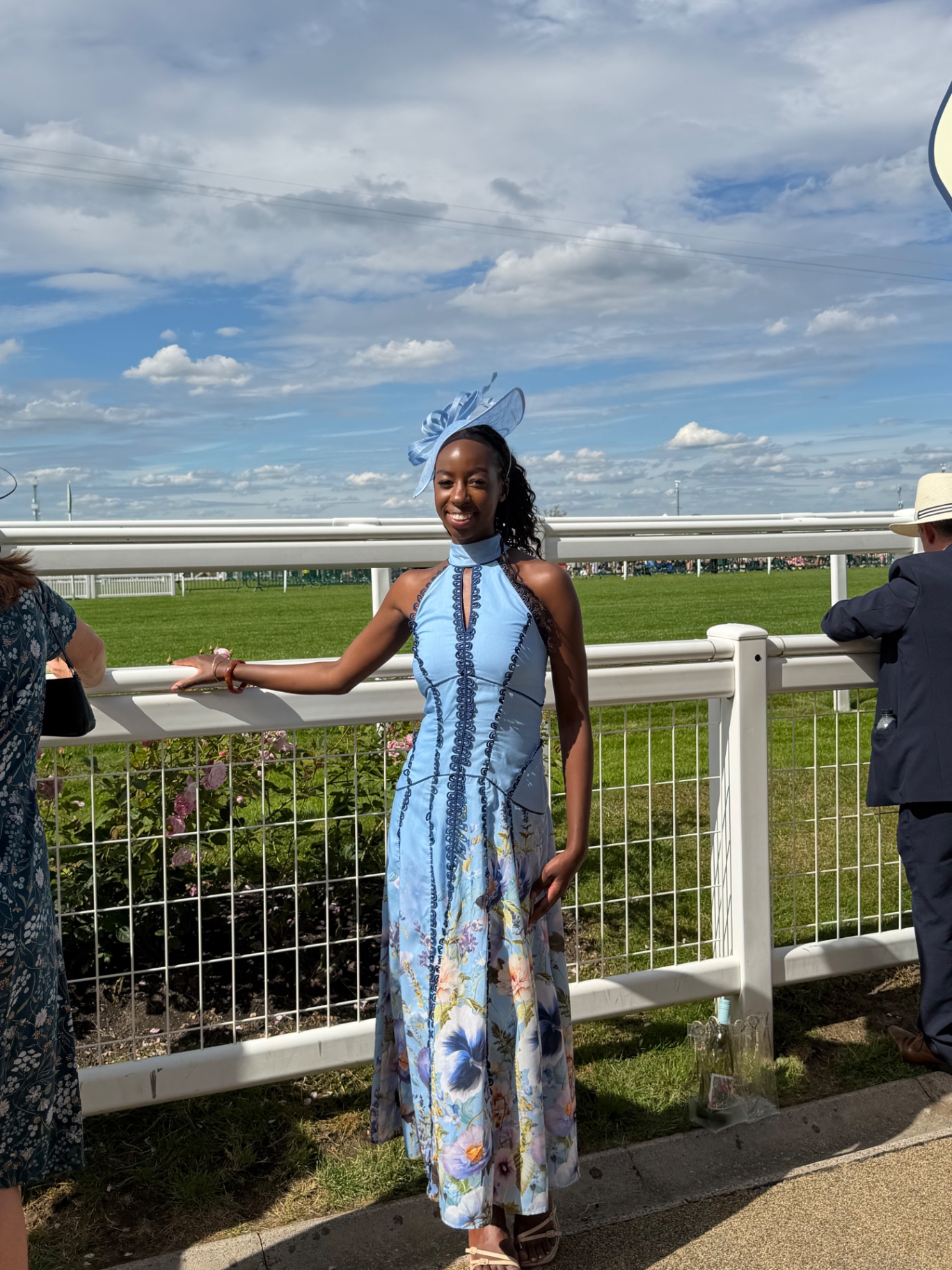 A member in a blue floral dress and fascinator by the racecourse rail
